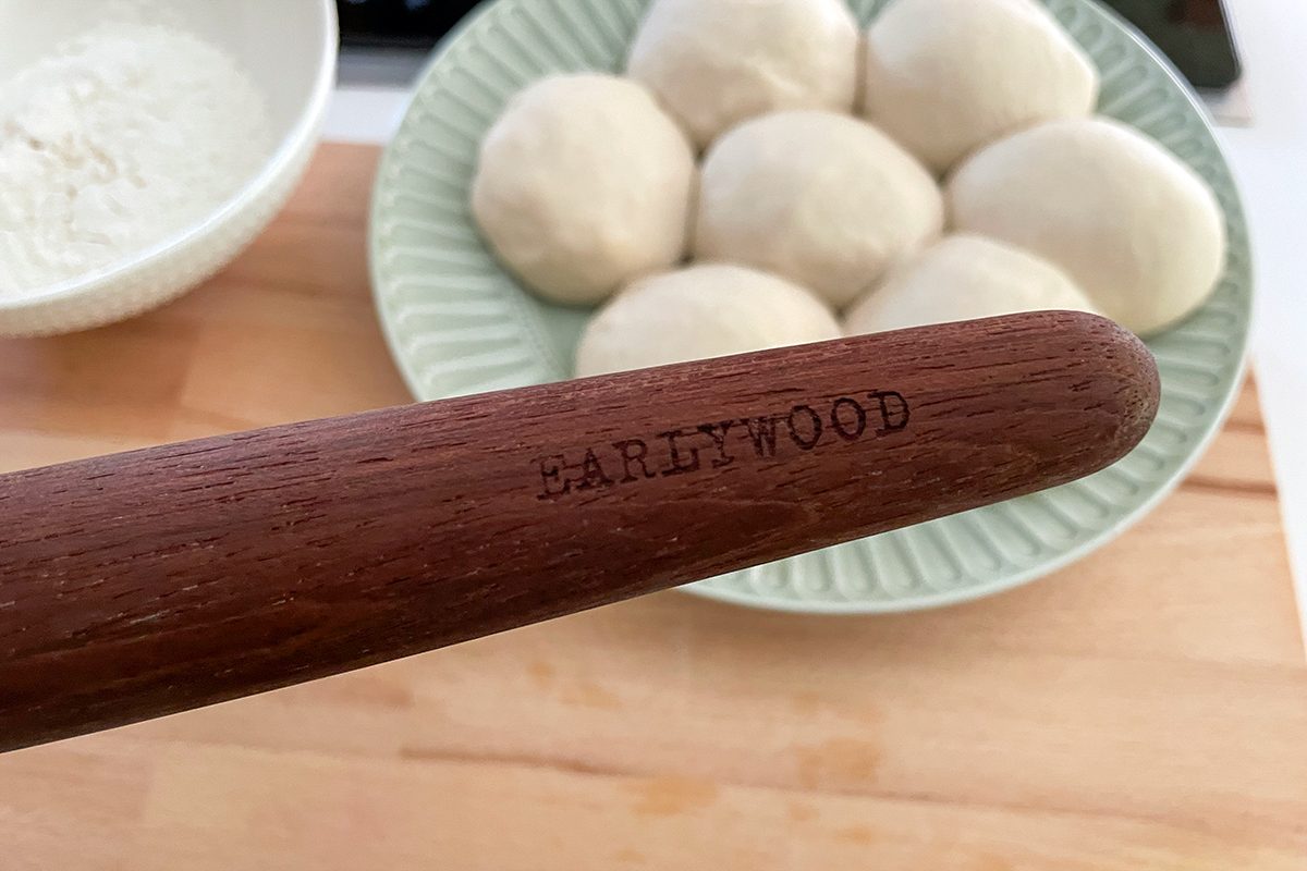 A wooden kitchen utensil engraved with "EARLYWOOD" is held in front of a plate with several balls of dough and a bowl of flour on a wooden surface.