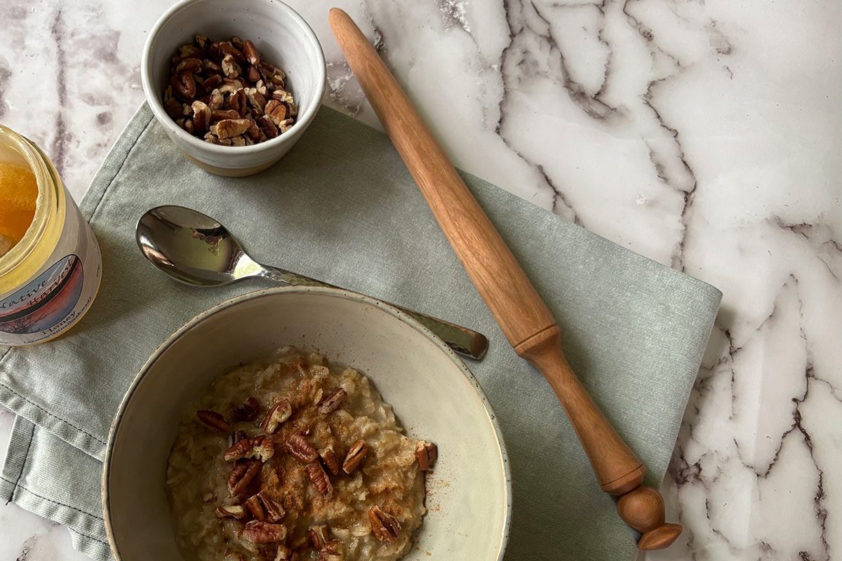 A bowl of oatmeal topped with chopped pecans sits on a folded green cloth next to a rolling pin, a spoon, a small bowl of pecans, and a jar of jam, all on a marble surface.