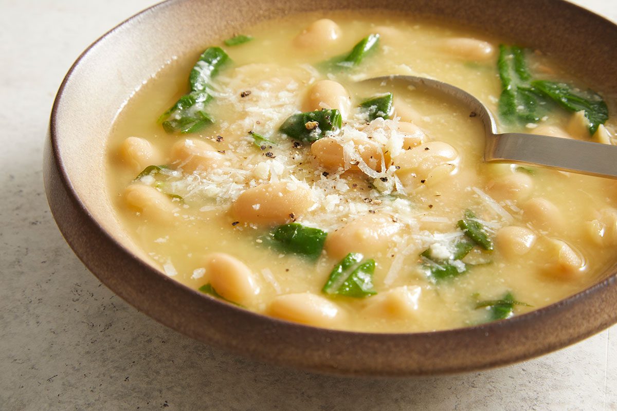 A bowl of creamy white bean soup with spinach, topped with grated cheese and black pepper, with a spoon resting in the soup.