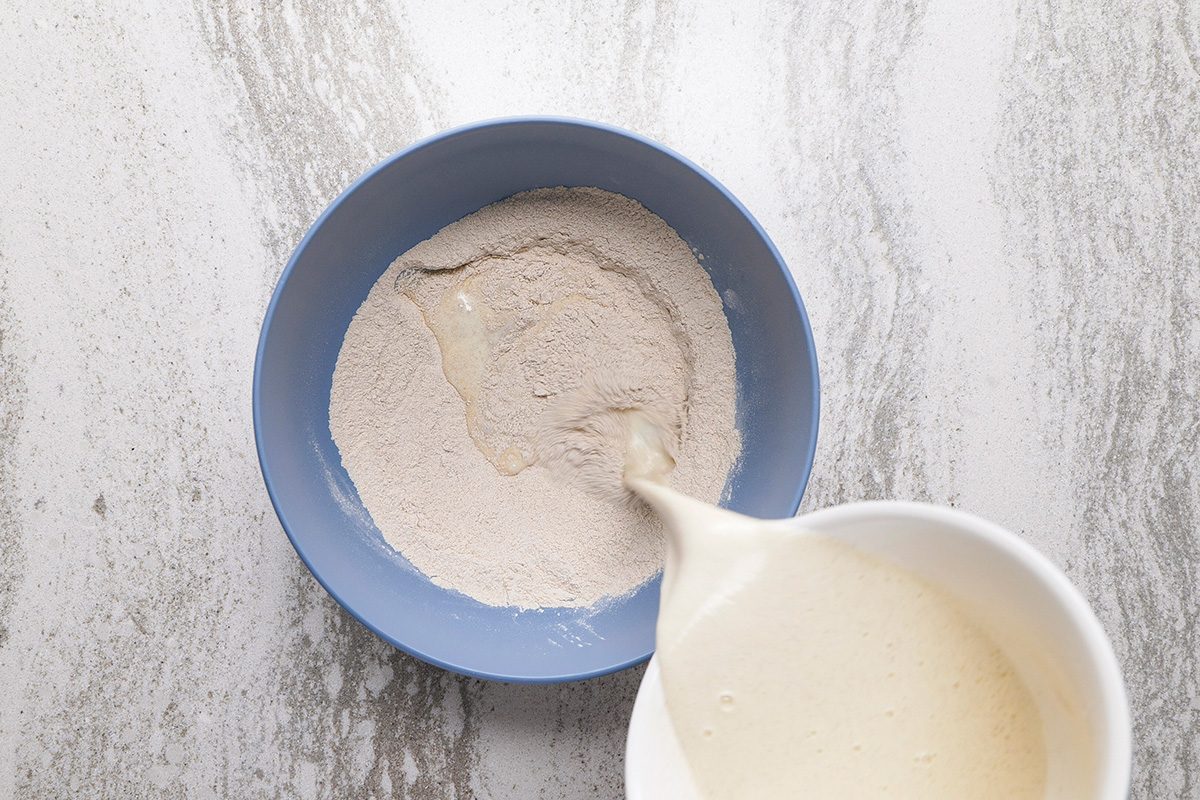 A blue bowl with flour and a well in the center sits on a marble surface, while a white pitcher pours liquid batter into the flour.