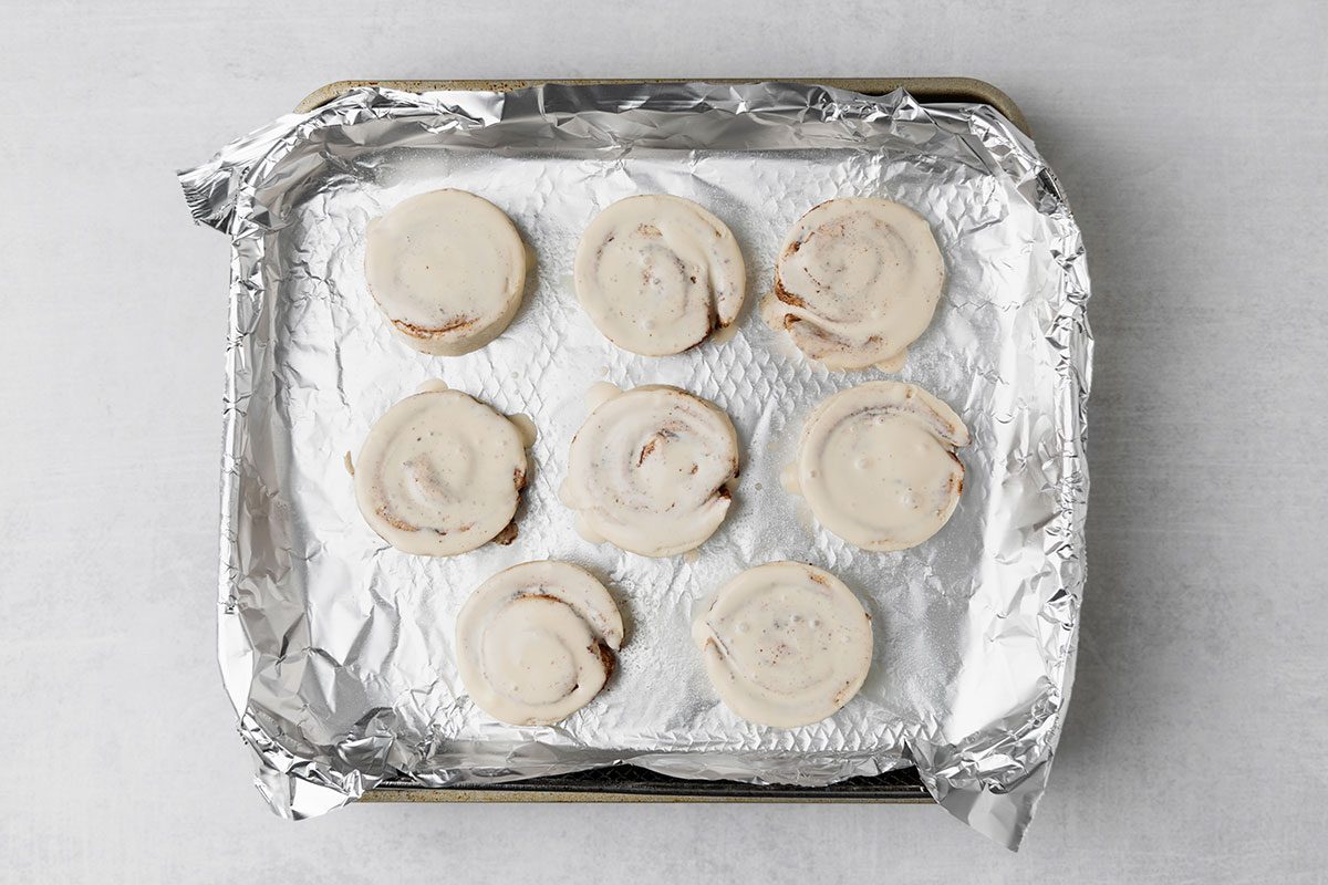 A baking tray lined with aluminum foil holds nine uncooked cinnamon rolls arranged in three rows. The rolls are spaced evenly apart and placed on a white surface.