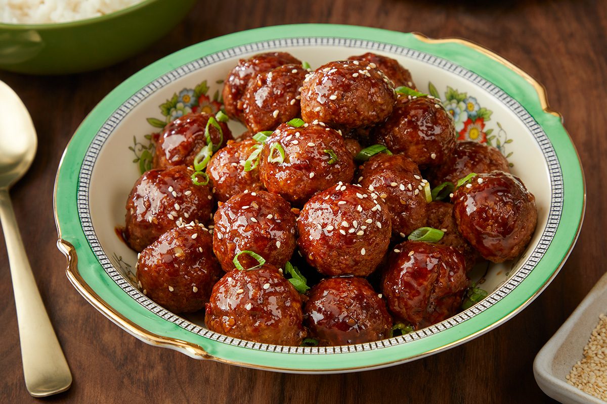 A decorative bowl filled with glazed meatballs garnished with sliced green onions and sprinkled with sesame seeds, placed on a wooden table.