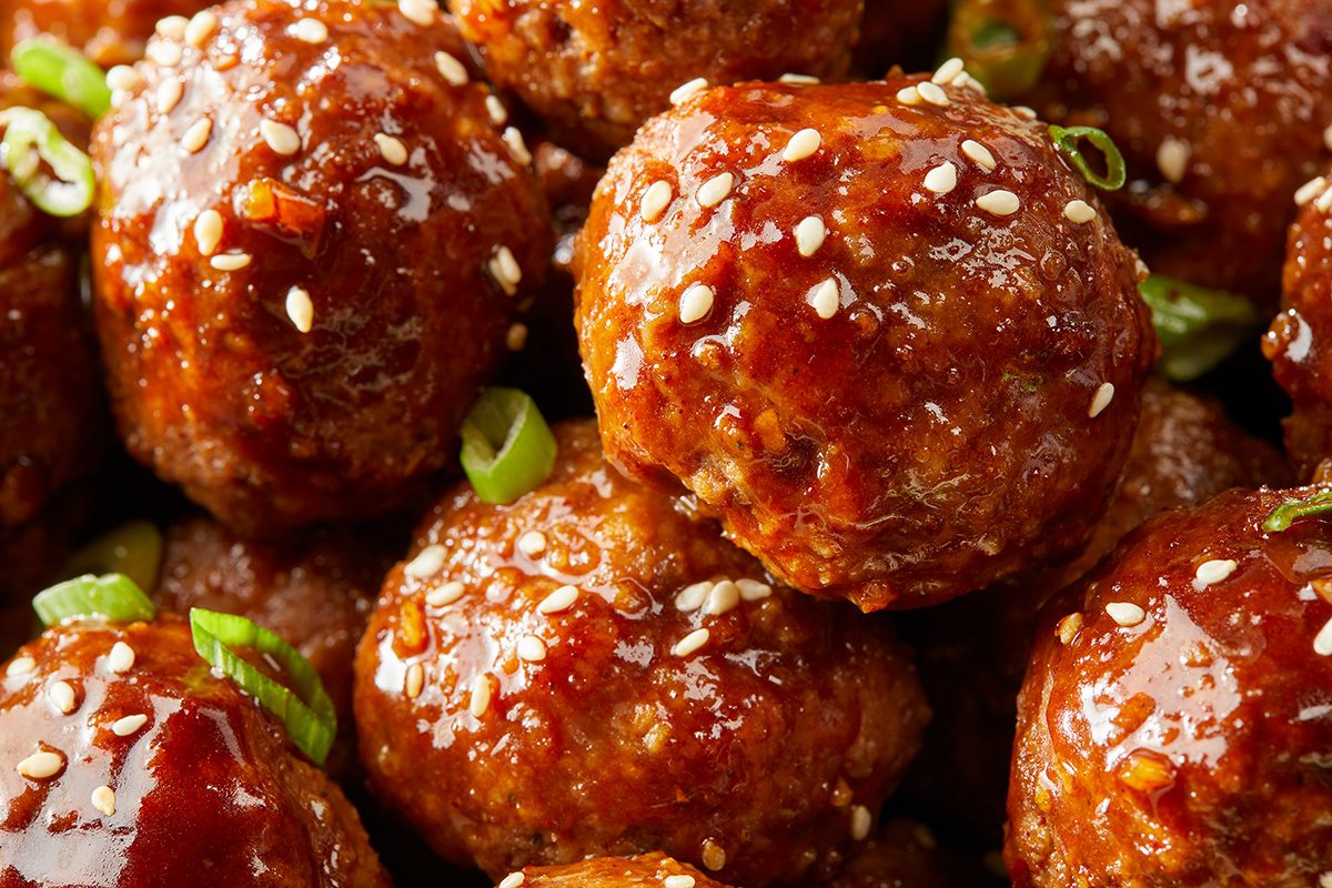 Glossy, sauce-covered meatballs are garnished with sesame seeds and sliced green onions, closely arranged in a pile.