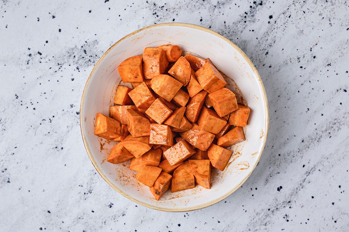 A white bowl filled with cubed sweet potatoes coated in spices, placed on a light speckled countertop.