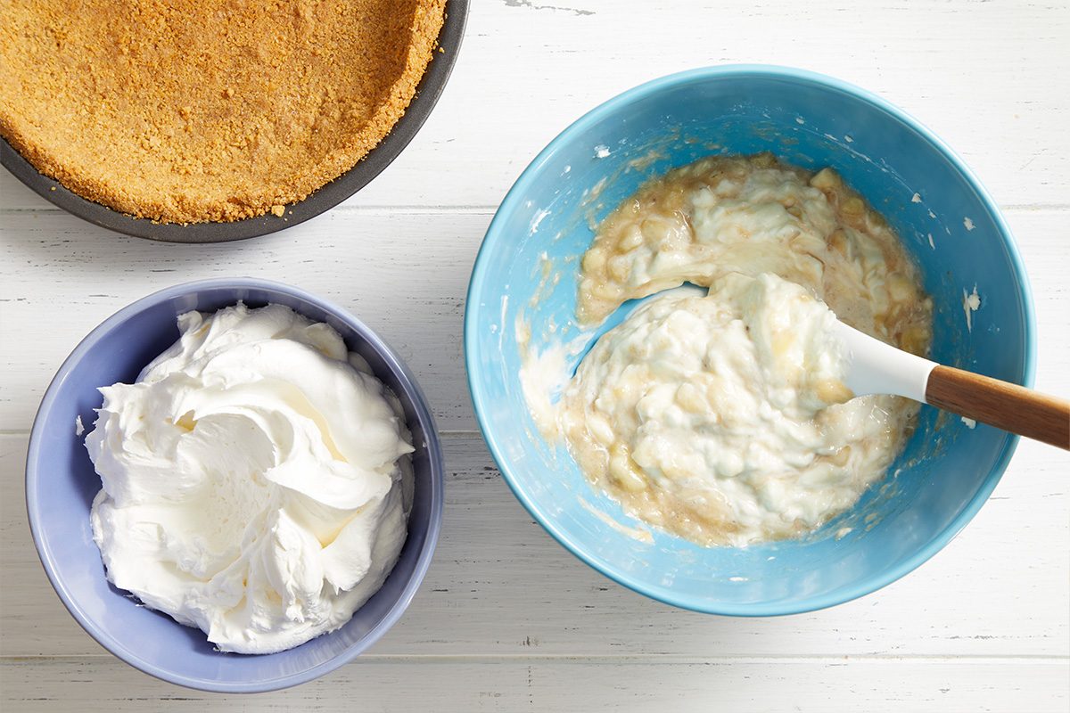 Two bowls and a pie crust on a white surface: one bowl with whipped cream, another with a creamy mixture and a spatula, and a pie crust in a pan.