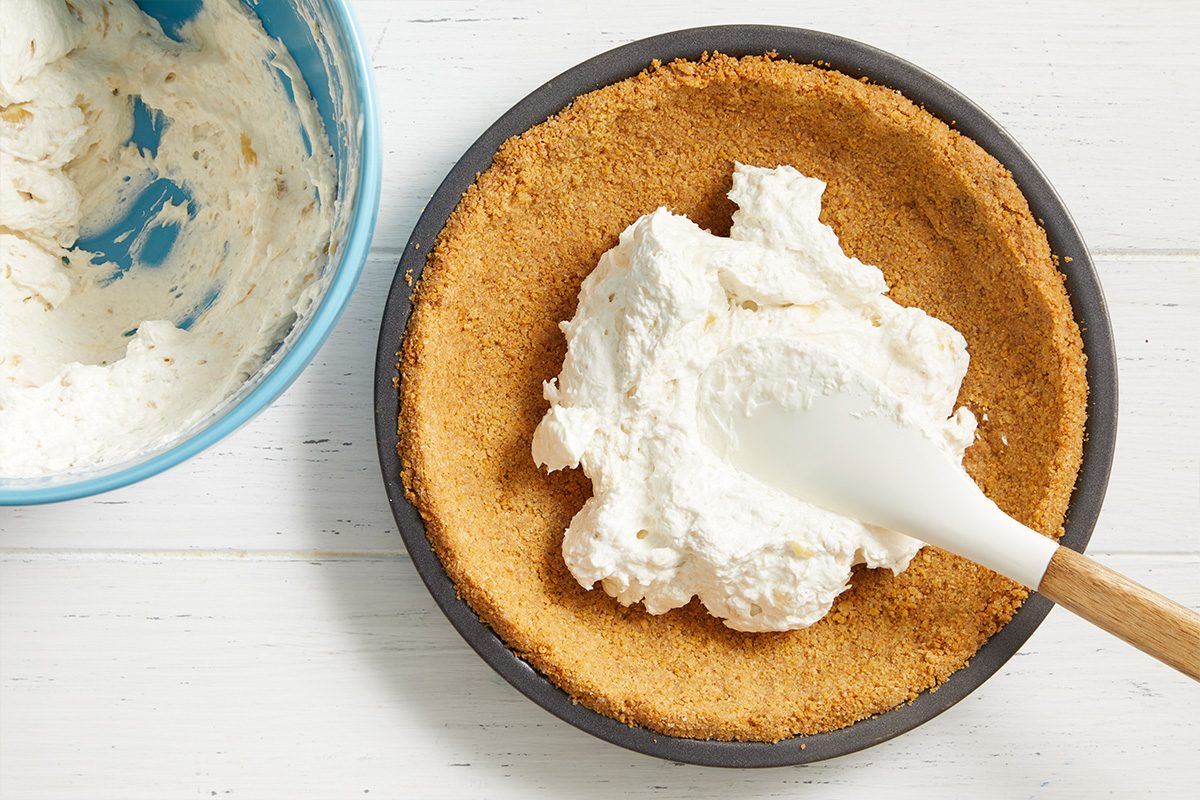 A pie crust in a round pan is being filled with a creamy white mixture using a spatula. A blue bowl with leftover filling is nearby on a white surface.
