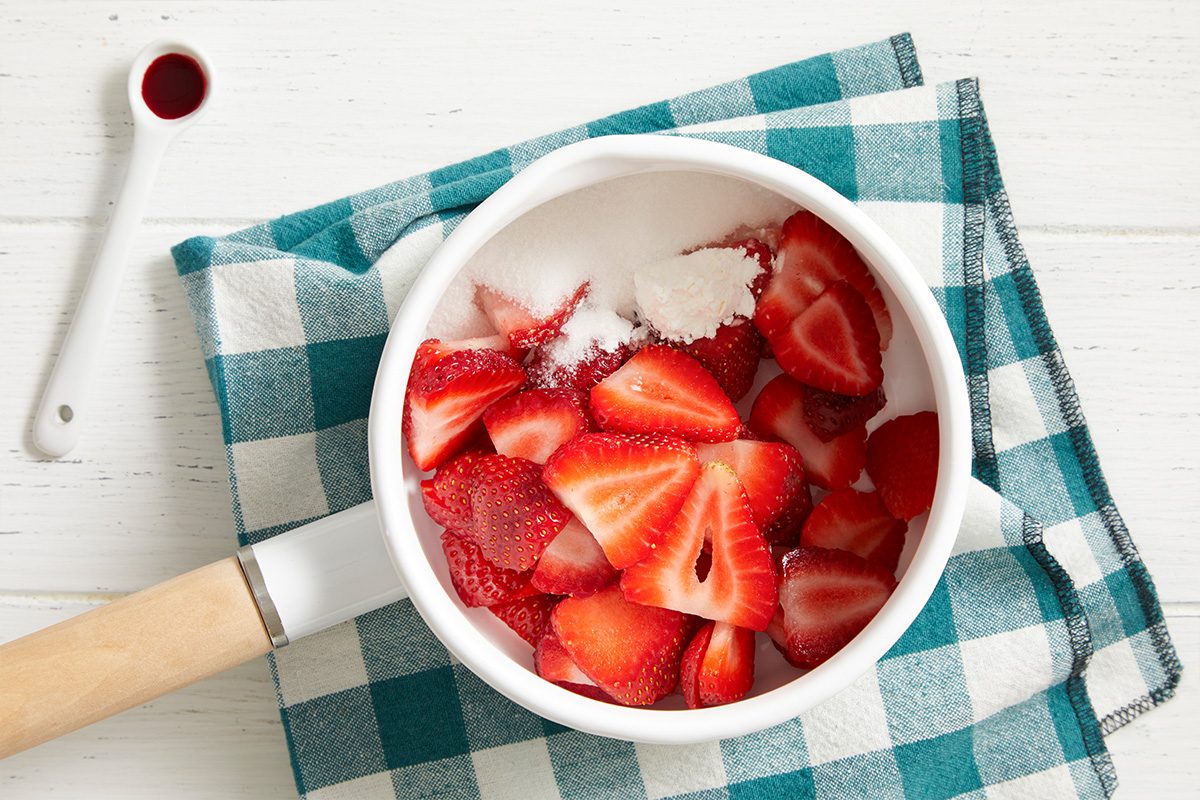 White saucepan with sliced strawberries, sugar, and cornstarch sits on a blue and white checkered cloth. A white measuring spoon with red liquid is nearby, all on a white wooden surface.