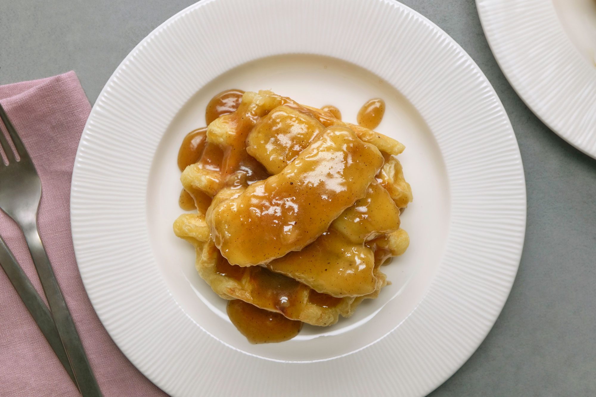 Overhead shot of Bananas Foster Sauce; Served warm with waffles; two forks resting on a napkin nearby; all set on a grey surface