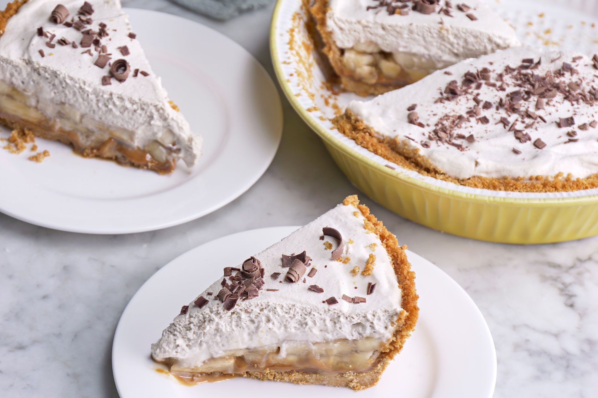 3/4 angle view shot of Banoffee Pie in a large pie plate; sliced and served on two plates; with a fork resting on a light green napkin; all set on a marble-textured surface