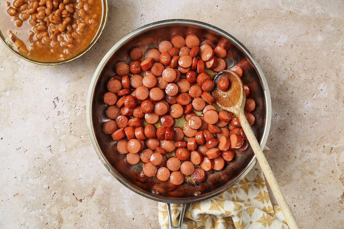 Overhead shot of a large skillet; melt butter over medium heat; Add hot dog slices and cook, stirring until browned; Then, add baked beans and heat through with a wooden spoon; A napkin lies on a marble surface;