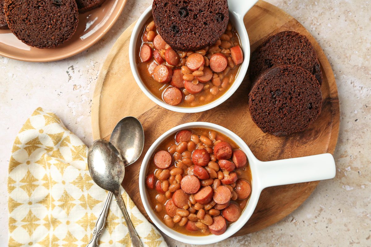 Overhead shot of Beanie Weenies in two white dishes with brown bread; served on a wooden round tray alongside two spoons and a napkin; all set on a marble surface;