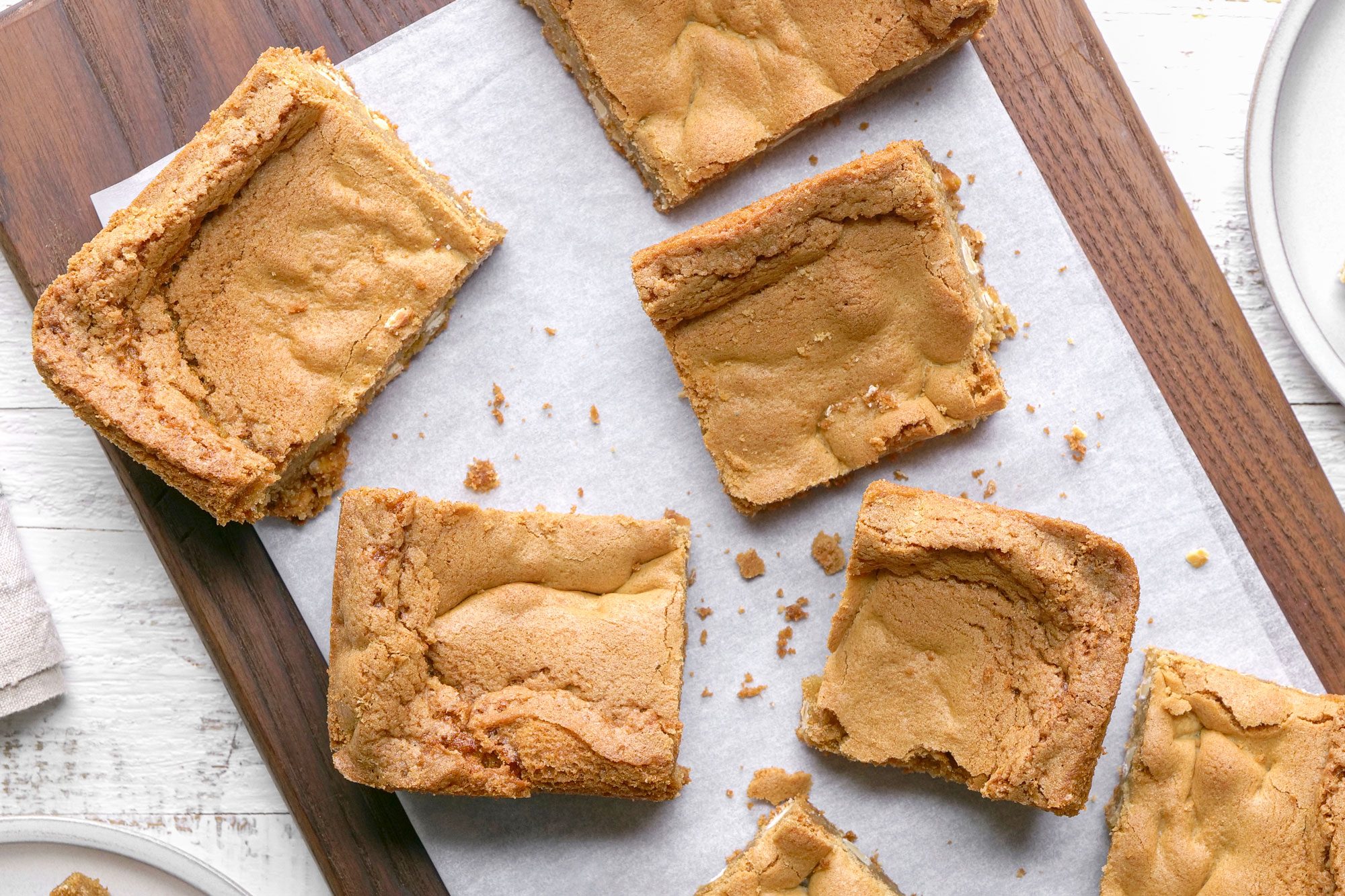 overhead shot of Squares of golden brown blondies are arranged on a parchment lined wooden cutting board with a few pieces on nearby white plates, all set on a white table