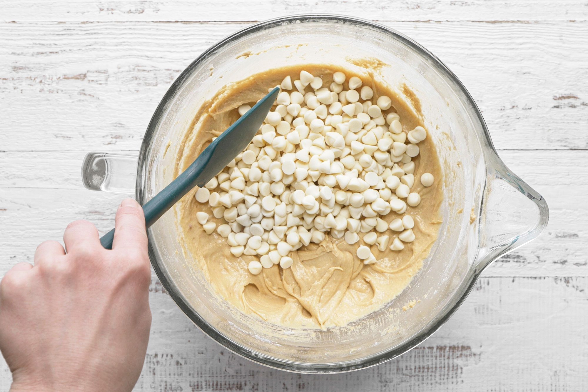 overhead shot of a hand holding a spatula mixes white chocolate chips into thick cookie dough in a clear glass bowl on a white wooden surface