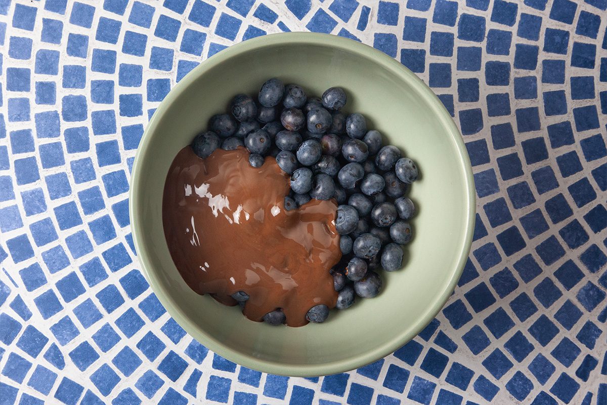 A bowl filled with fresh blueberries and chocolate sauce on one side, placed on a blue and white mosaic-tiled surface.