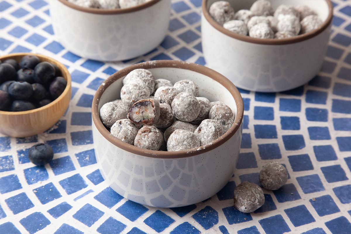Small white bowls filled with blueberries coated in powdered sugar on a blue and white patterned surface. A wooden bowl with fresh blueberries is also visible.