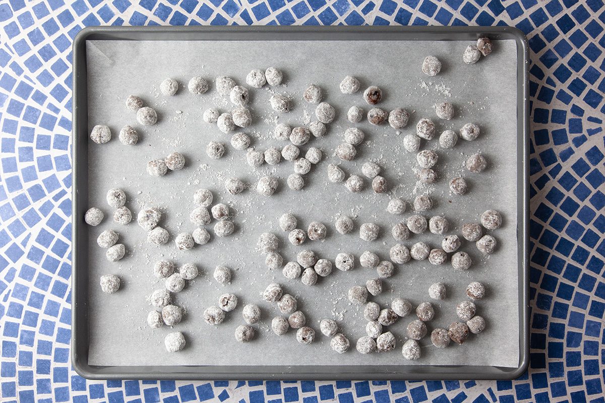 A baking sheet lined with parchment paper holds many small, round, powdered sugar-coated balls, possibly cookie dough, resting on a blue and white mosaic-patterned surface.