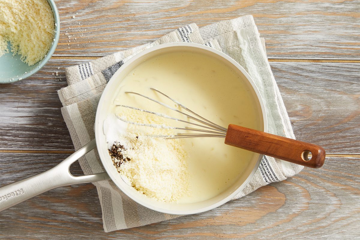 A saucepan filled with creamy white sauce sits on a cloth-covered wooden surface. A whisk with a wooden handle rests inside, and grated cheese and seasoning are being added to the sauce.