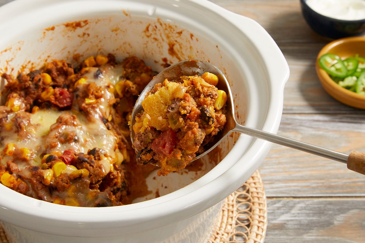 A close-up of a crockpot filled with cheesy ground beef, corn, beans, and tomatoes, with a large serving spoon lifting out a portion. The crockpot sits on a wooden table next to small bowls of toppings.