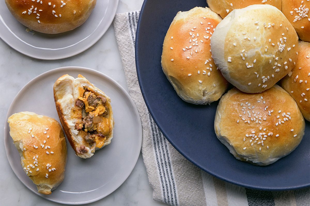 A plate of golden brown, sesame-topped baked buns sits next to a plate with one whole bun and one bun split open, revealing a savory meat and cheese filling inside.