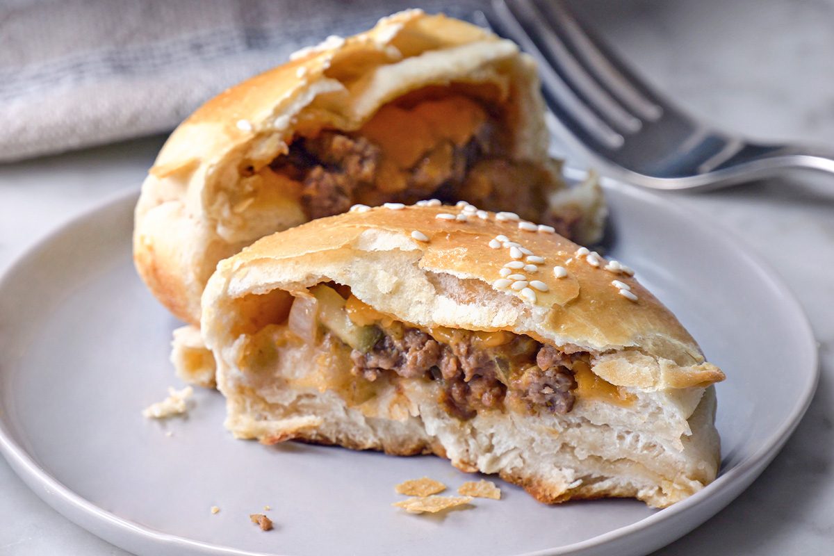 A stuffed bread roll cut in half on a gray plate, showing a filling of ground beef, onions, and melted cheese. A fork rests in the background. The roll is topped with sesame seeds.