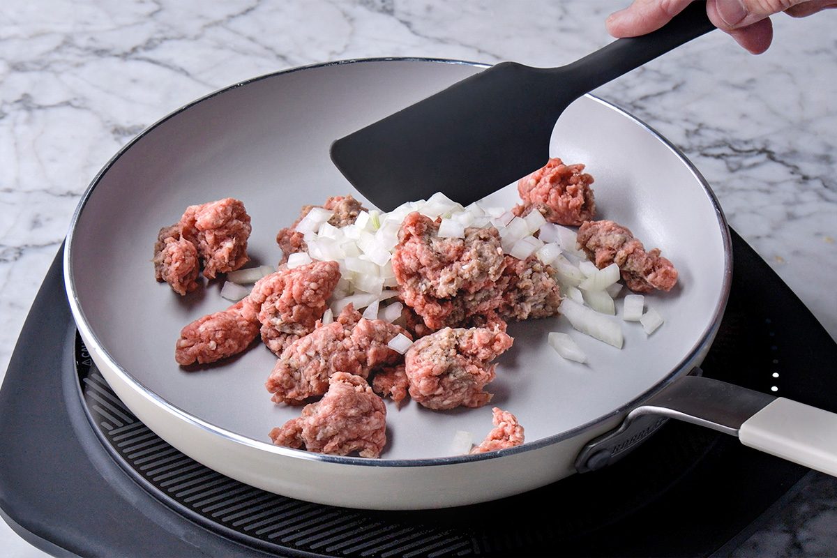 Ground beef and chopped onions being stirred in a white frying pan with a black spatula on a stovetop, against a marble countertop background.
