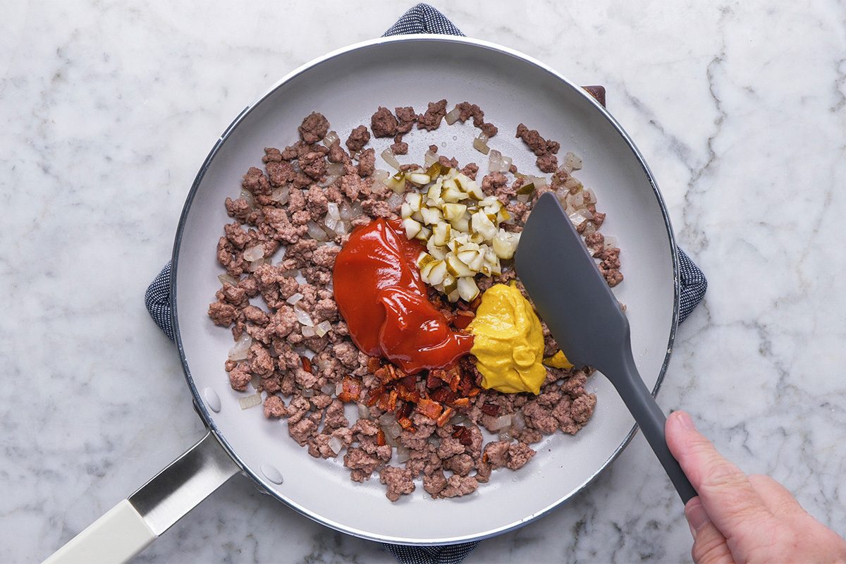 A hand holds a spatula over a skillet with cooked ground beef, chopped onions, ketchup, mustard, and chopped pickles on a marble countertop.
