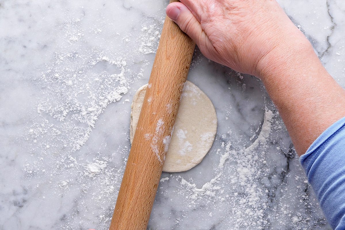 A hand using a wooden rolling pin to flatten a small round piece of dough on a floured marble surface.