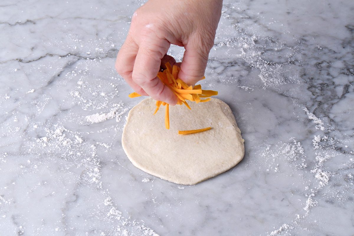 A hand sprinkles shredded cheddar cheese onto a small, round piece of dough on a floured marble surface.