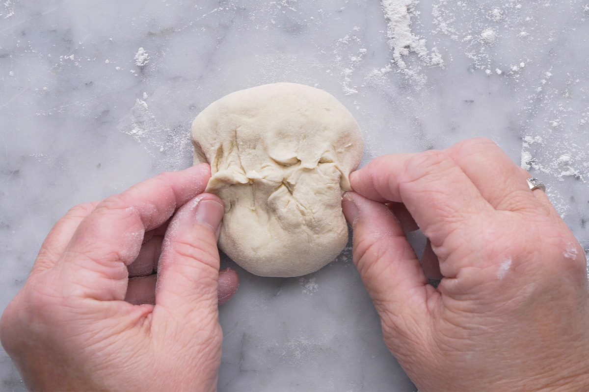 Two hands are shaping a small ball of dough on a lightly floured surface, pinching the dough together at the top.