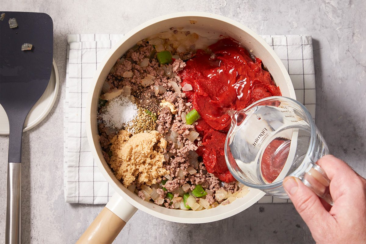 A saucepan on a checkered cloth contains ground meat, tomato paste, chopped onions, green peppers, seasonings, and brown sugar, while a hand pours water into the pan. A spatula is nearby.