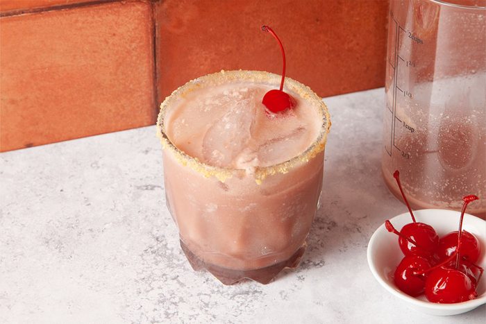 A creamy pink cocktail in a glass with a sugared rim, garnished with a cherry on top. Next to it is a small bowl containing several maraschino cherries, all set against a light countertop and brick background.