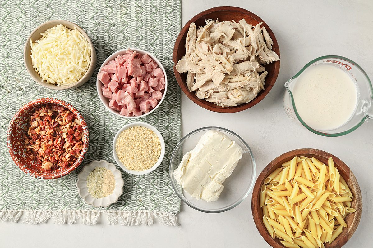 Various ingredients are arranged on a light surface, including shredded cheese, diced ham, shredded chicken, cream, cooked pasta, cream cheese, bread crumbs, and a bowl of diced tomatoes with herbs.