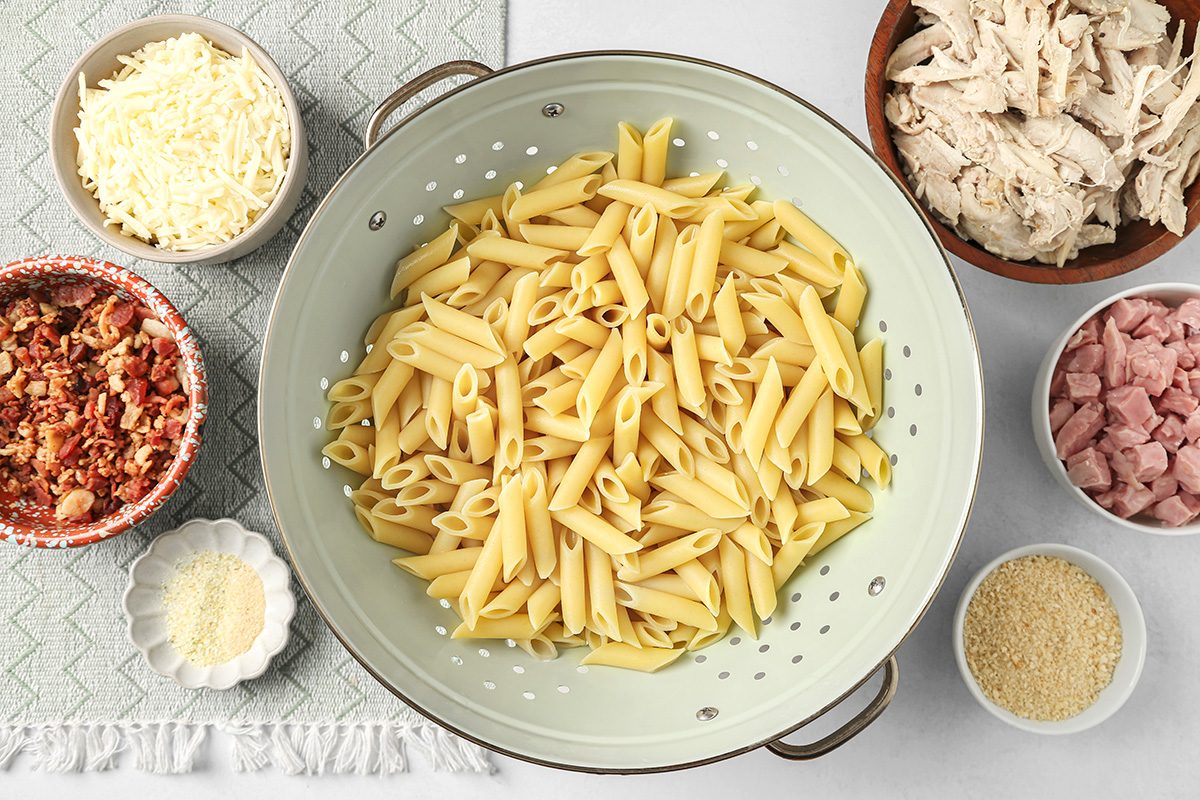 Cooked penne pasta in a colander, surrounded by bowls containing shredded cheese, cooked bacon bits, shredded chicken, diced ham, and grated Parmesan cheese, arranged on a light surface.
