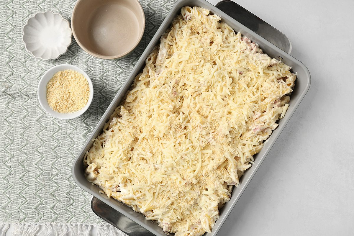 A rectangular baking dish filled with an uncooked pasta casserole topped with shredded cheese, next to two empty bowls and a small bowl of grated cheese on a textured, light-colored surface.