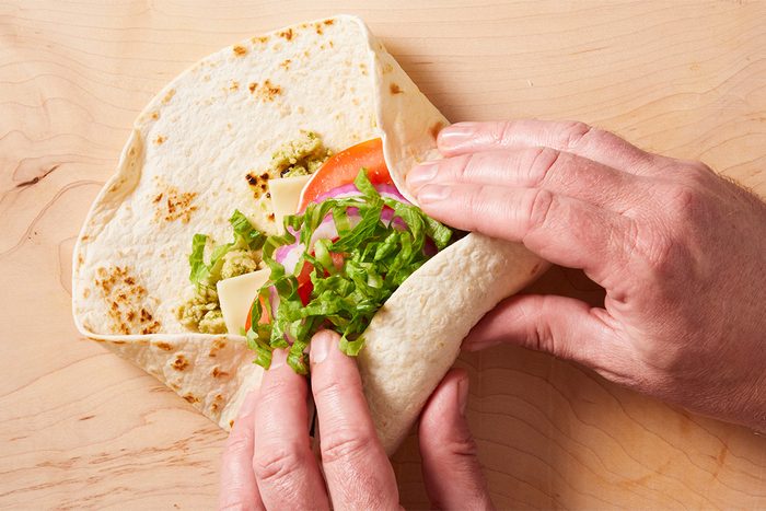 Hands folding a tortilla wrap filled with lettuce, sliced tomato, red onion, cheese, and guacamole on a light wooden surface.