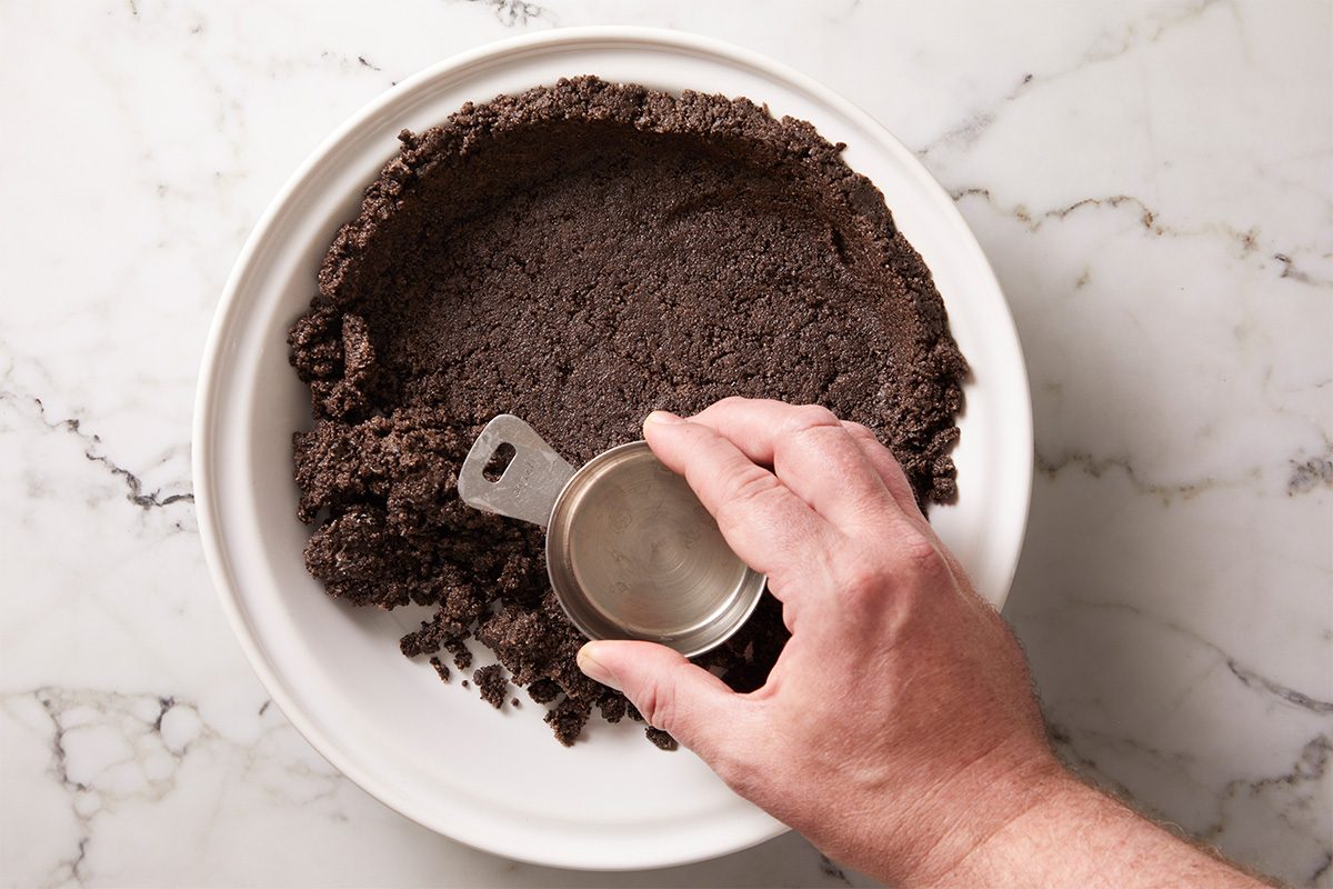 A hand presses an Oreo cookie crumb mixture into a pie dish using a metal measuring cup to form a crust on a white marble surface.