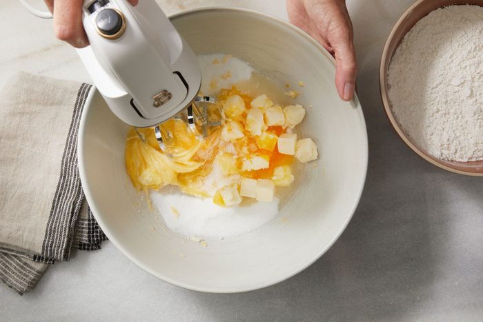 overhead shot of a person uses a hand mixer to blend butter, sugar, and eggs in a bowl, with a bowl of flour and a striped kitchen towel nearby on a marble countertop