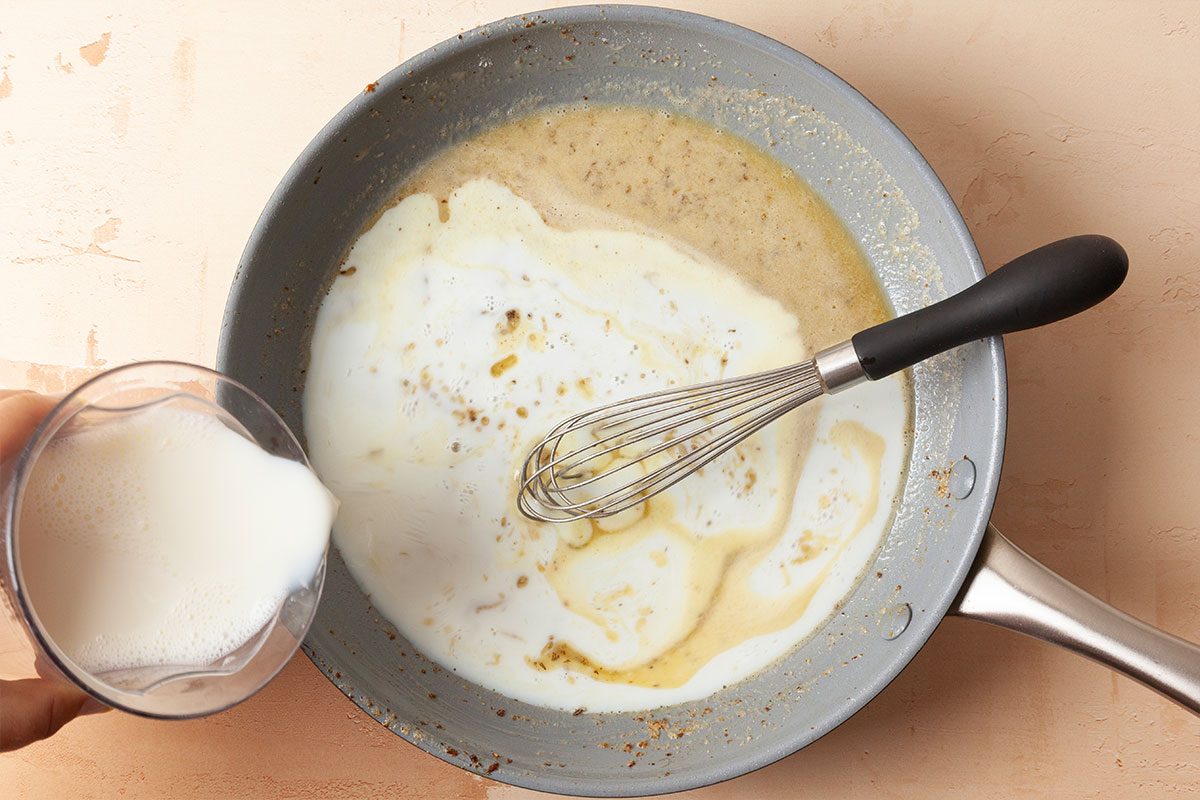 A hand pours milk from a glass into a saucepan with a creamy mixture, while a whisk rests in the pan, ready to stir the ingredients together.