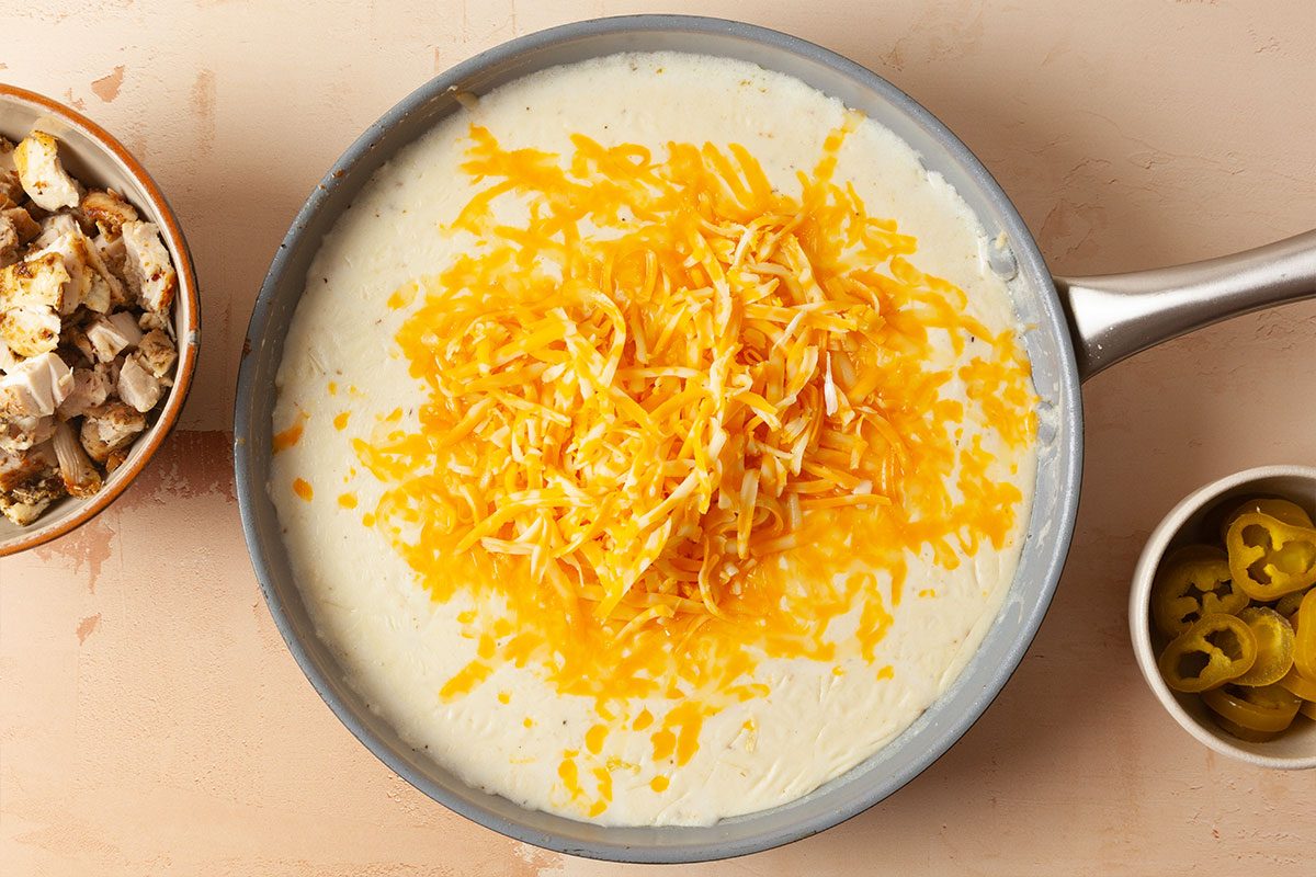 A skillet filled with creamy white sauce topped with shredded cheddar cheese sits on a counter. Nearby are bowls of cooked chicken pieces and sliced jalapeños.