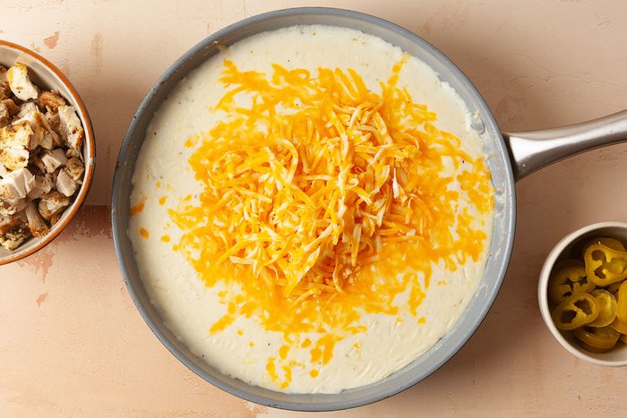 A skillet filled with creamy white sauce topped with shredded cheddar cheese sits on a counter. Nearby are bowls of cooked chicken pieces and sliced jalapeños.