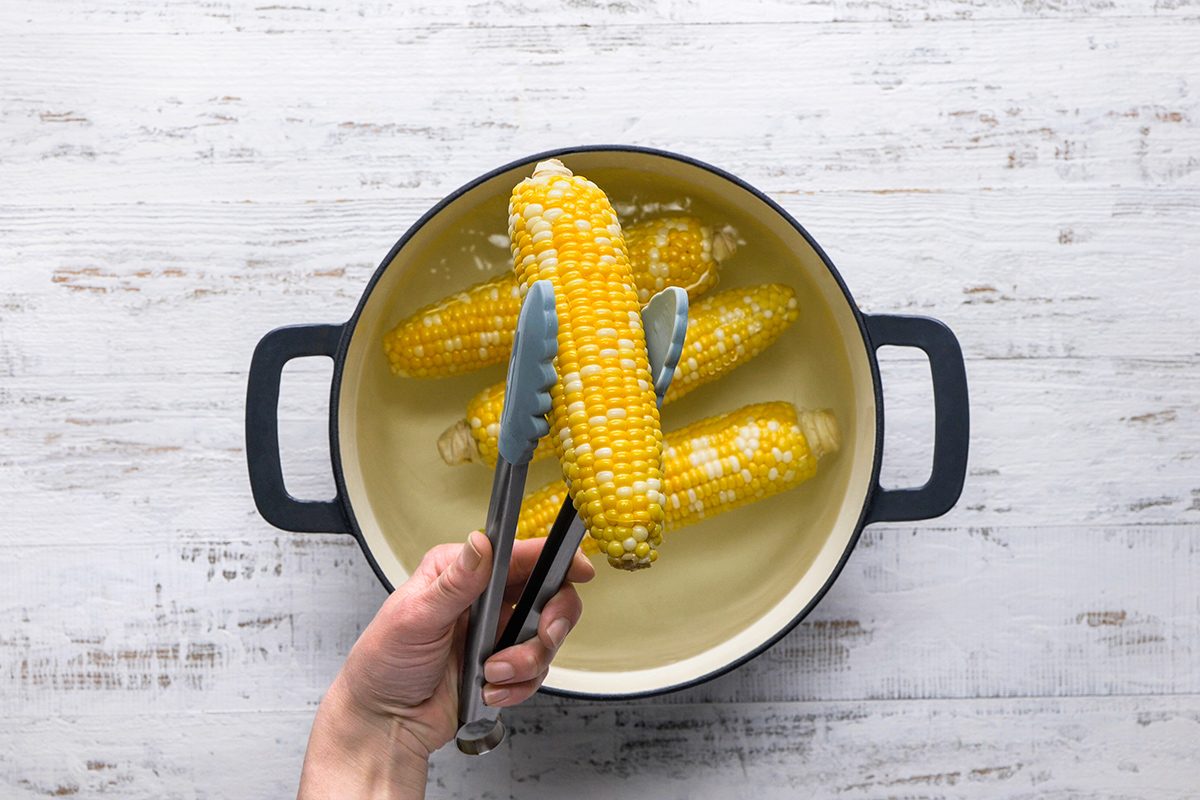 A hand uses metal tongs to hold two ears of cooked corn over a pot of water, with more corn still in the pot, on a white wooden surface.