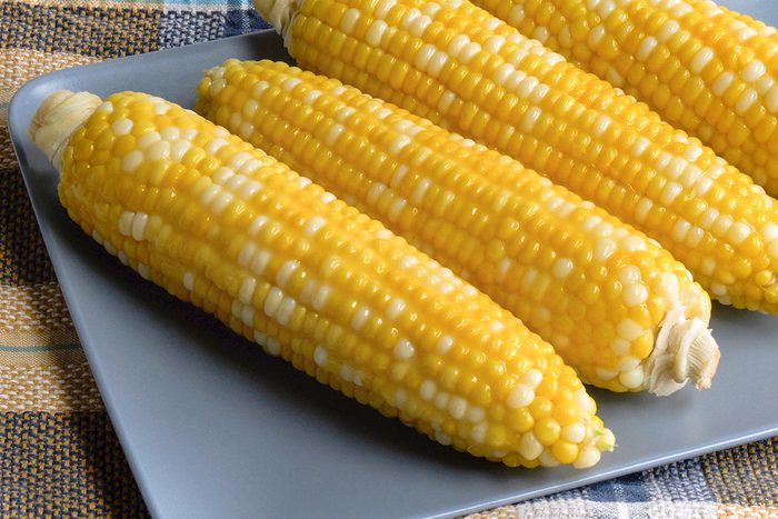 Three ears of cooked corn on the cob with yellow and white kernels are arranged side by side on a square gray plate, set on a plaid-patterned tablecloth.
