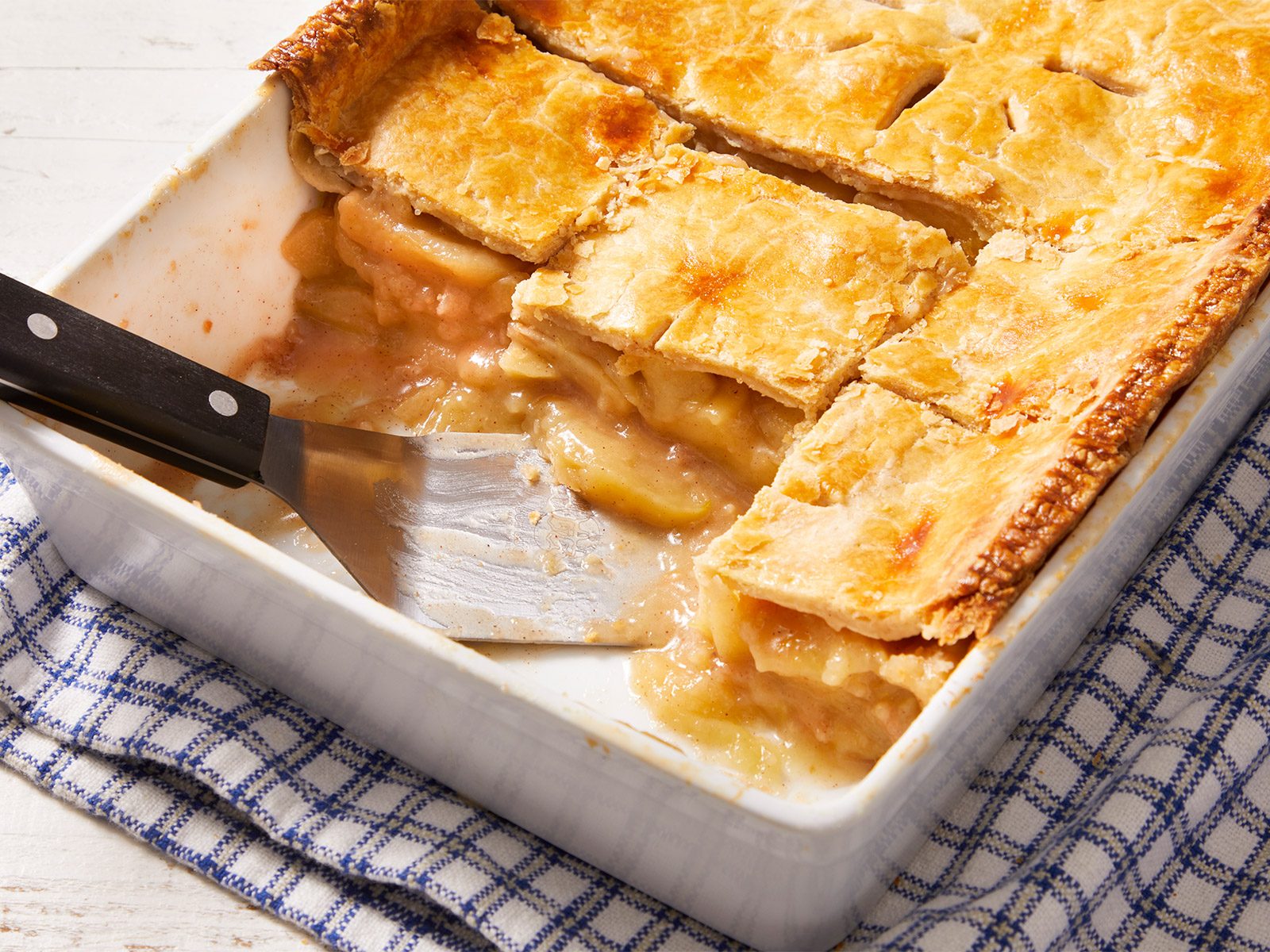 3/4 shot of Deep-Dish Apple Pie with a spatula serving it in a white tray kept on a checkered kitchen cloth.
