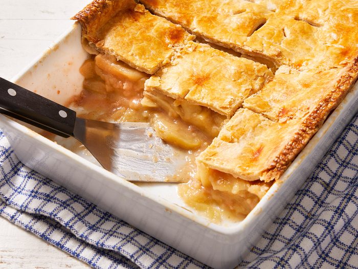 3/4 shot of Deep-Dish Apple Pie with a spatula serving it in a white tray kept on a checkered kitchen cloth.
