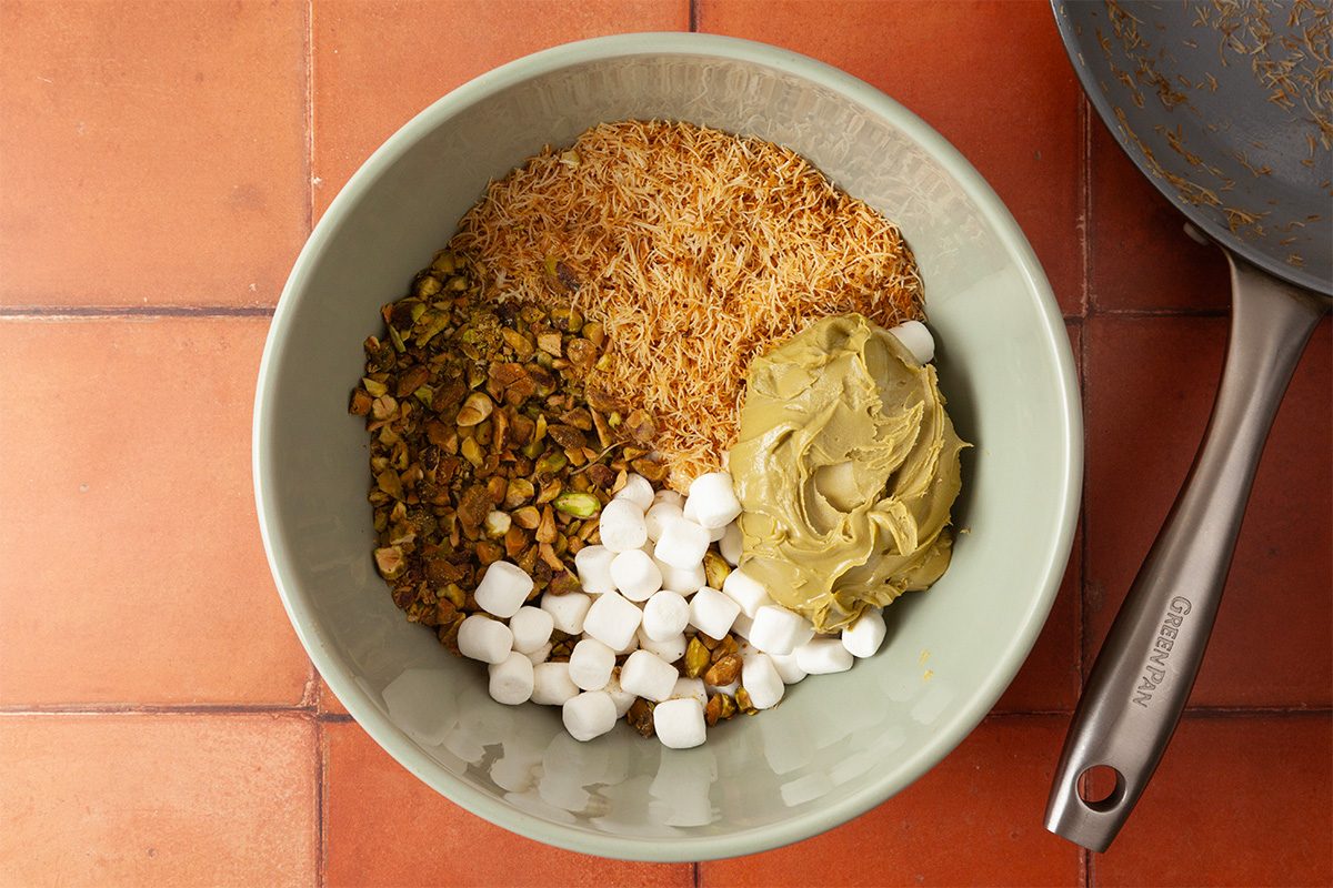 A bowl holds shredded toasted coconut, chopped pistachios, mini marshmallows, and a spoonful of green pistachio paste. A frying pan is partially visible beside the bowl on a tiled countertop.