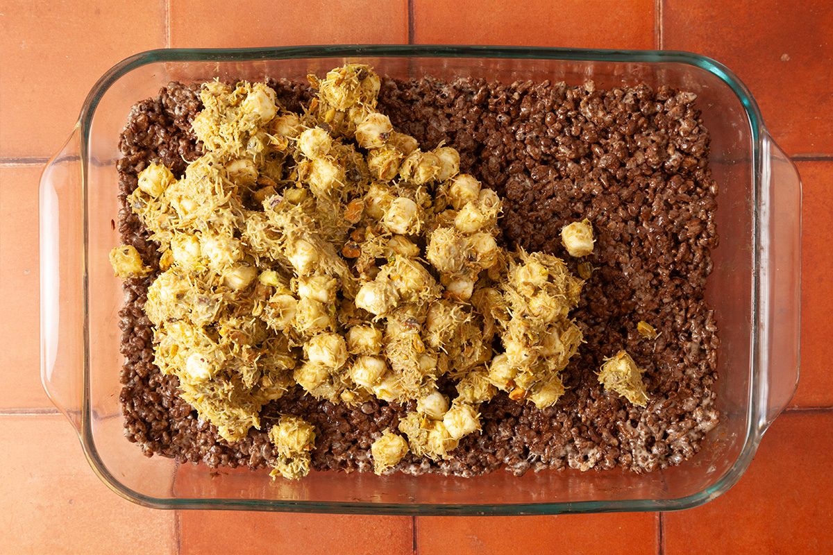 A glass baking dish filled with a layer of chocolate rice cereal topped with clusters of what appears to be coated popcorn or cereal, on an orange tiled surface.