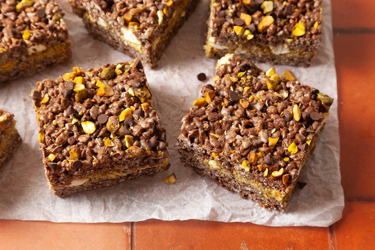 Chocolate and nut cereal bars cut into squares are arranged on white parchment paper over a tiled surface. The bars have a crumbly texture with visible chocolate chips and pieces of nuts on top.