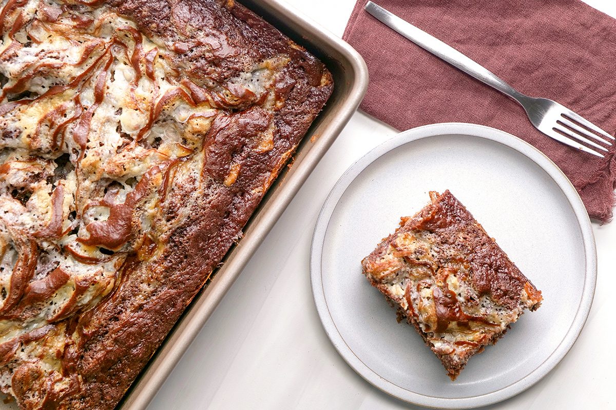 A baking tray with marbled cheesecake brownies and a single square brownie served on a white plate beside a fork and brown napkin.