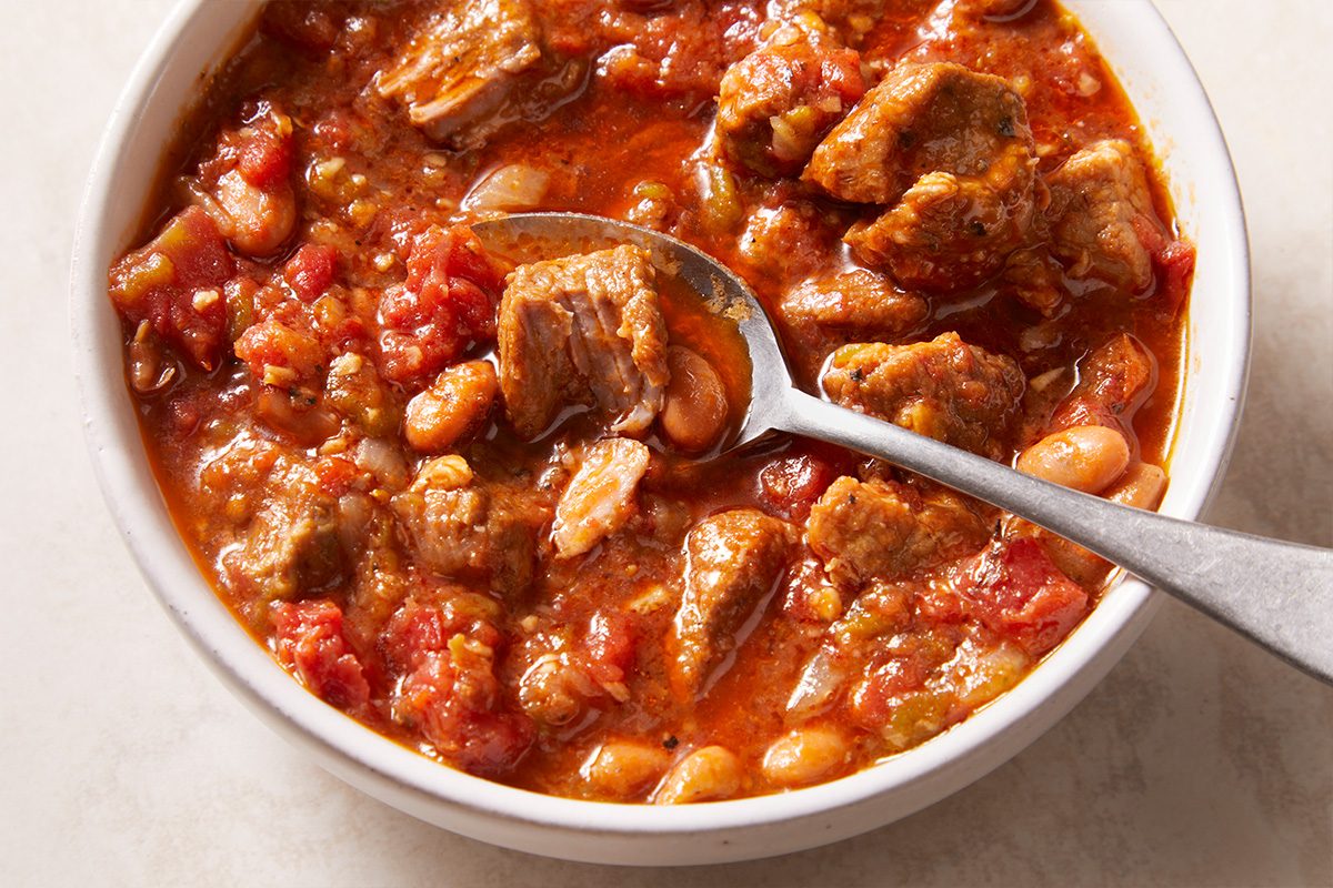 A bowl of chunky chili filled with beef, beans, and diced tomatoes, with a metal spoon resting inside the bowl.