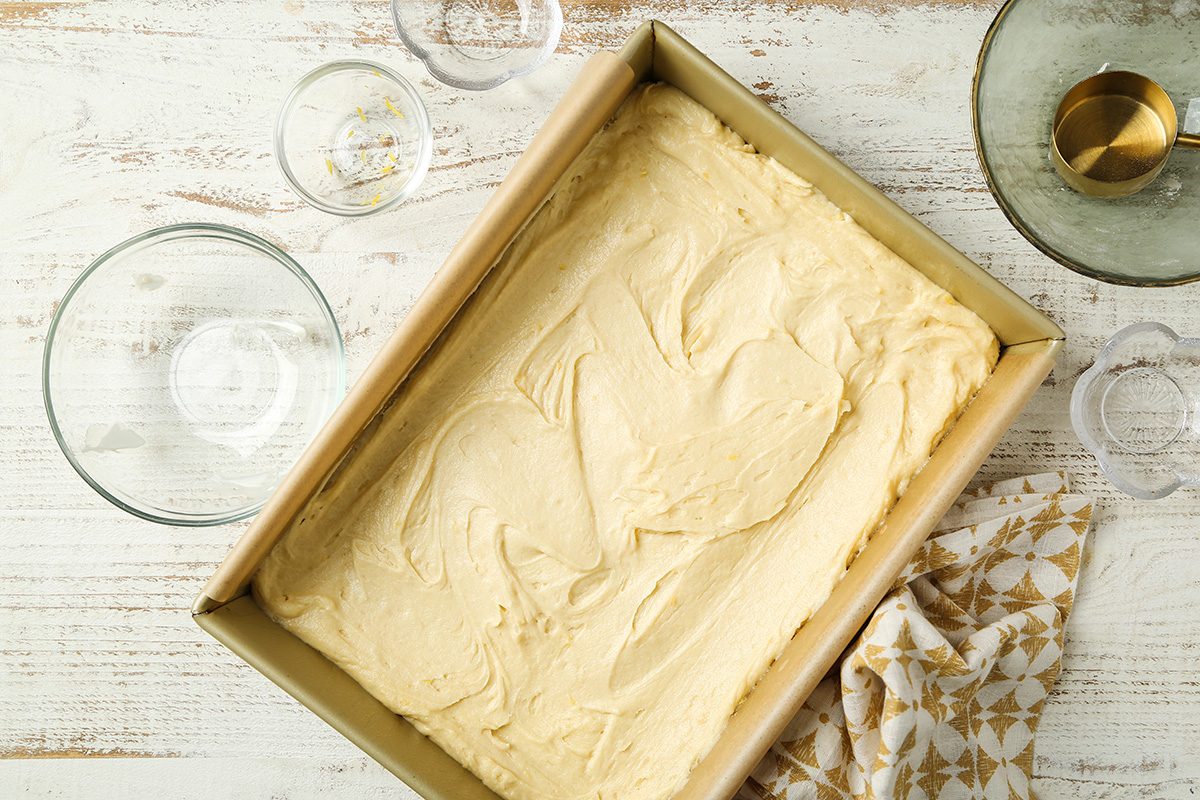 A rectangular baking pan filled with an even layer of cake batter sits on a white wooden surface surrounded by empty glass bowls and a patterned kitchen towel.
